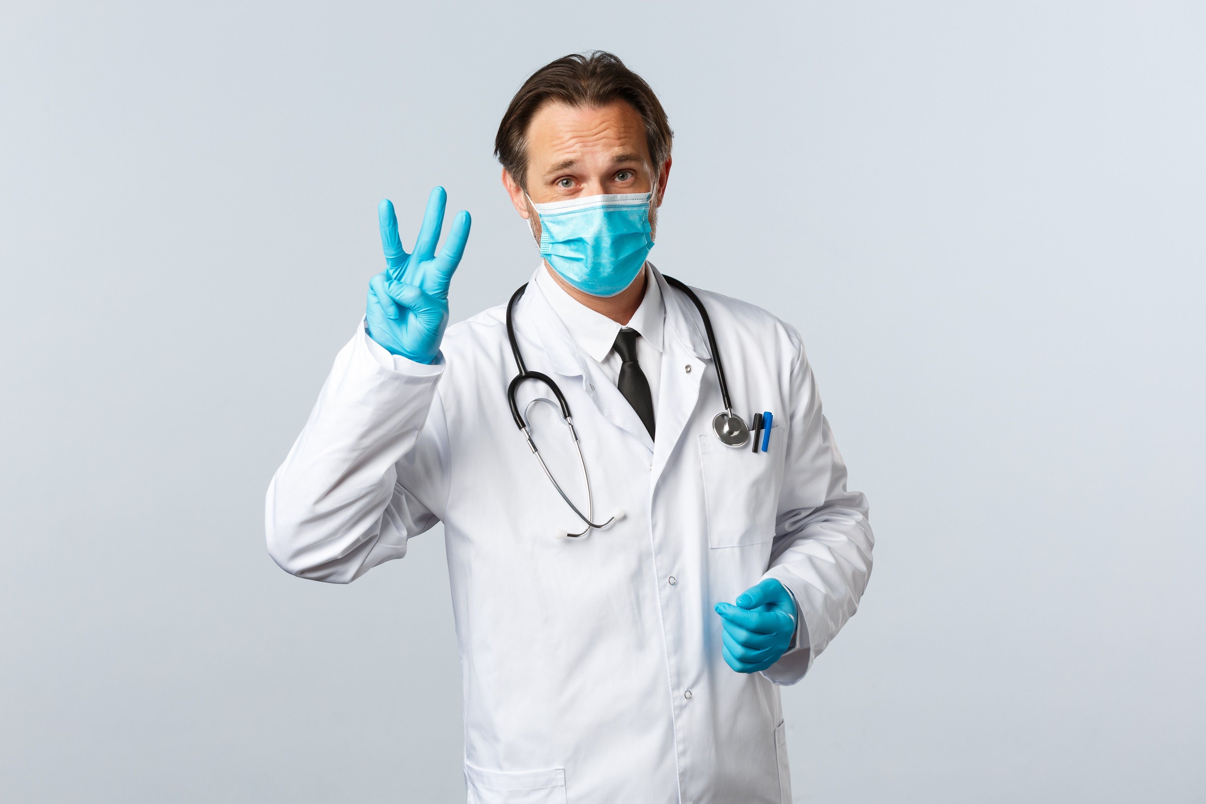 Male urologist in blue gloves and mask holding up three fingers to indicate the three prostate zones. 